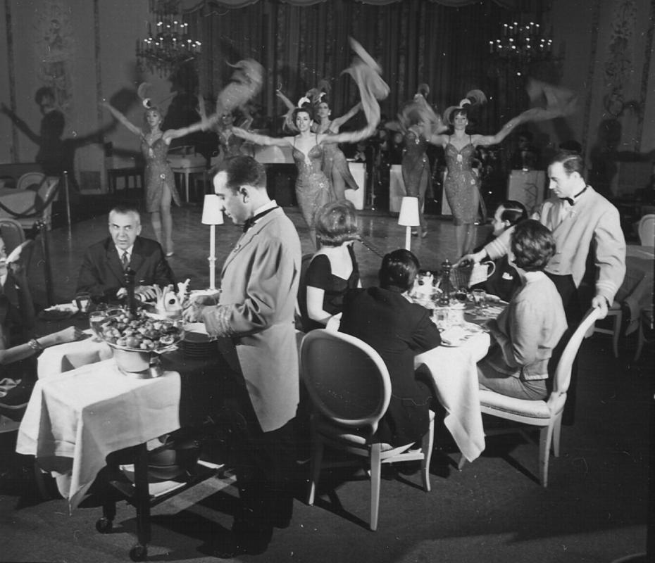 Patrons dining at a cabaret in mid-century Montréal while showgirls in feathered costumes perform on stage, with chandeliers and elegant decor in the background.