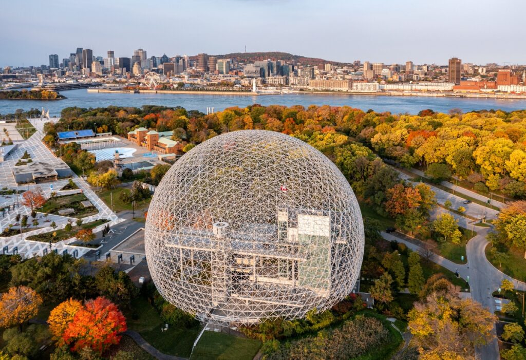 Aerial view of the Montreal Biosphere surrounded by vibrant fall foliage on Île Sainte-Hélène, with the downtown skyline and Mount Royal in the background.