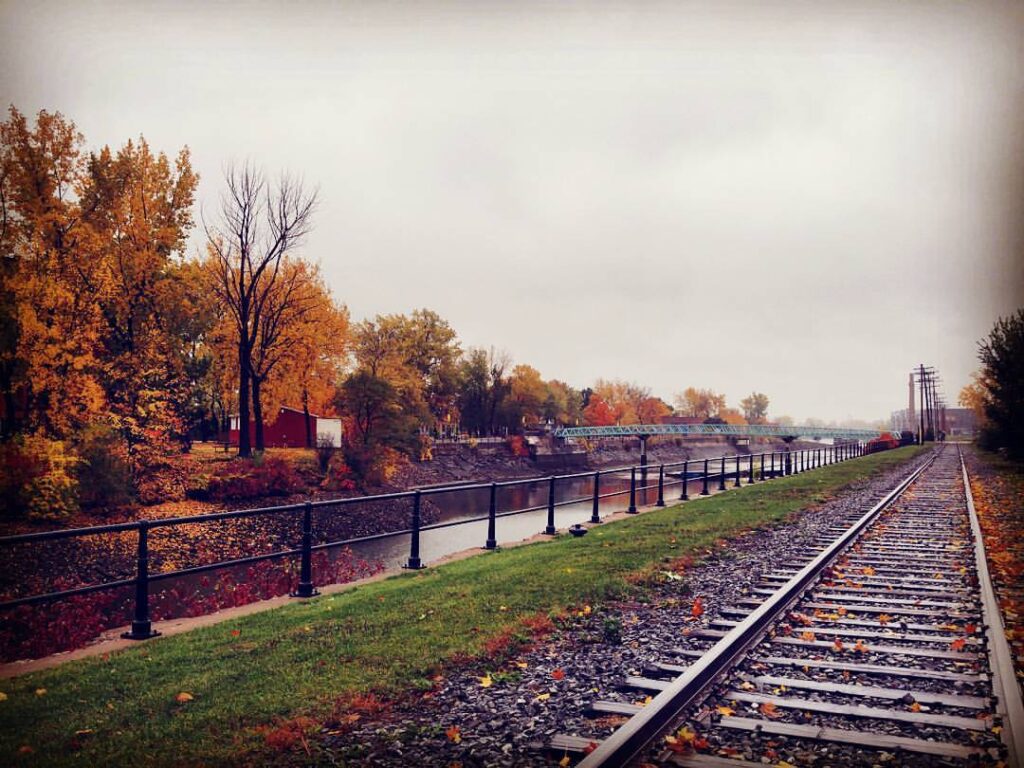Railway tracks beside the Lachine Canal in Montreal during fall, with orange and yellow foliage lining the canal on a misty autumn day.